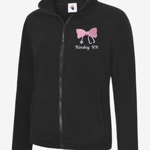 May include: Black fleece jacket with a full zip and a high collar. The jacket features a pink bow and stethoscope design with the text "Kinsley RN" embroidered on the chest. It has two front pockets.