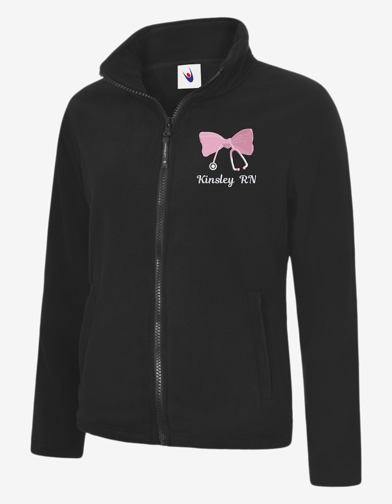 May include: Black fleece jacket with a full zip and a high collar. The jacket features a pink bow and stethoscope design with the text "Kinsley RN" embroidered on the chest. It has two front pockets.