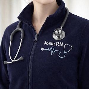 May include: Navy blue fleece zip-up jacket with a stethoscope around the neck. The jacket has the embroidered text "Josie, RN" and a light blue stylized EKG and stethoscope design. A medical professional's attire.