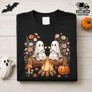 Op de afbeelding: Een zwart t-shirt met een afbeelding van twee witte geesten die op een boomstam zitten en marshmallows roosteren boven een kampvuur. Het ontwerp bevat kleurrijke wilde bloemen, een pompoen en paddenstoelen voor een herfsttafereel.