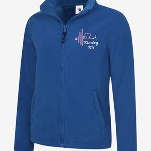 May include: Royal blue fleece jacket with a full zip, high collar, and two front pockets. The jacket features a pink heartbeat design with a heart and the text "Kinsley RN". A comfortable and warm outerwear option.