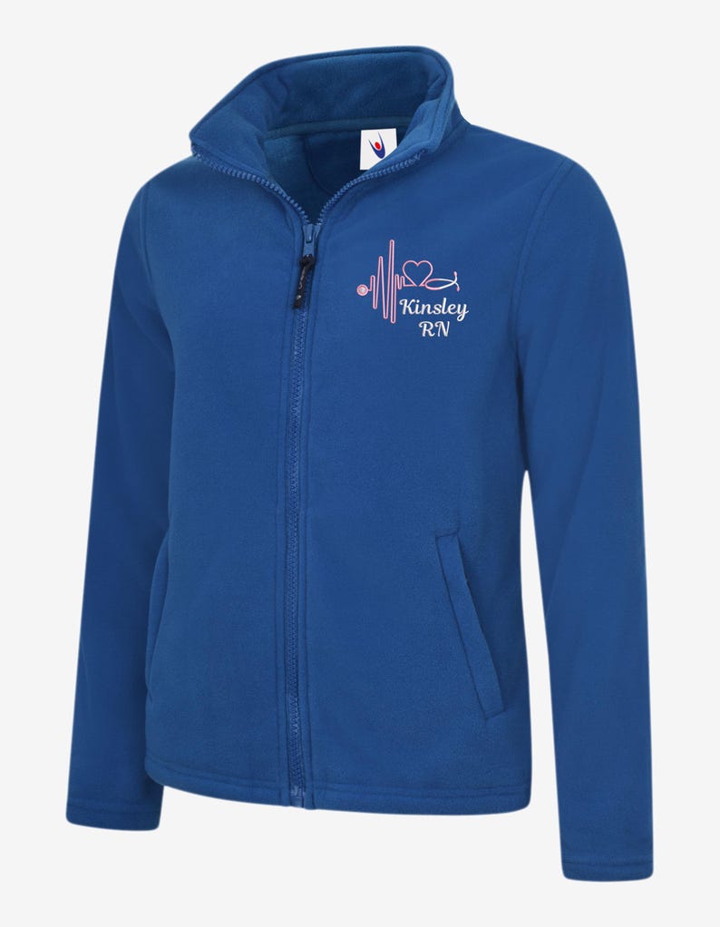 May include: Royal blue fleece jacket with a full zip, high collar, and two front pockets. The jacket features a pink heartbeat design with a heart and the text "Kinsley RN". A comfortable and warm outerwear option.