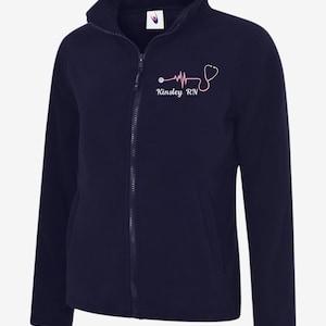 May include: Navy blue fleece jacket with a full zip and a high collar. A pink stethoscope and heartbeat design is printed on the left chest, along with the name "Kinsley RN". The jacket has two front pockets.
