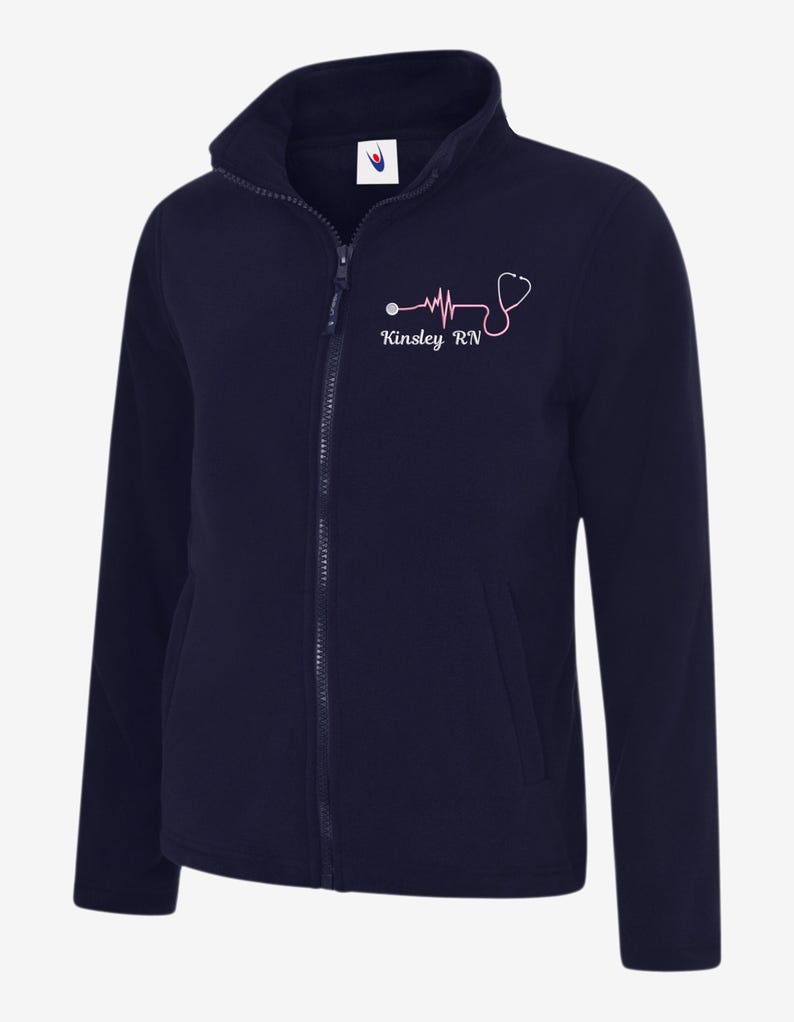 May include: Navy blue fleece jacket with a full zip and a high collar. A pink stethoscope and heartbeat design is printed on the left chest, along with the name "Kinsley RN". The jacket has two front pockets.