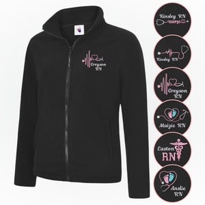 May include: A black fleece zip-up jacket featuring a pink embroidered design on the left chest with a heartbeat line, a heart, a stethoscope, and the text Greyson RN. Six circular patches display various nurse-themed designs and names.