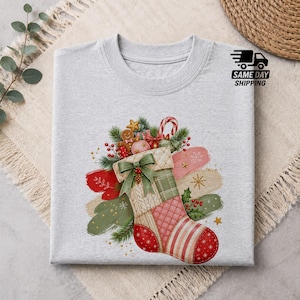 Puede incluir: Una camiseta gris claro con un gráfico de un calcetín navideño decorado con un lazo verde, ramas de pino, bayas y un hombre de jengibre. El calcetín tiene un patrón acolchado y rayas rojas y blancas. Se ve el texto SAME DAY SHIPPING.