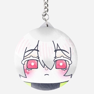 May include: A round keychain featuring a cartoon character with white hair, red eyes, and a sad expression. The character has pink blush and is wearing a black and green outfit. The keychain is attached to a silver chain.