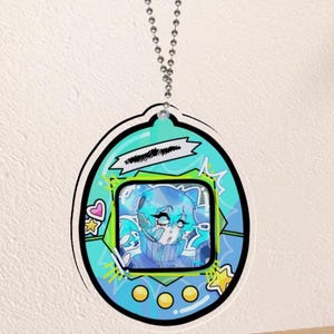 May include: A teal and black acrylic charm in the shape of a retro digital pet device. It features a cartoon illustration of a blue-haired character. The charm is suspended from a silver ball chain.