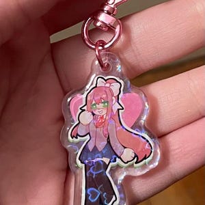 Puede incluir: Un llavero rosa en forma de corazón con un diseño de personaje de dibujos animados. El personaje tiene cabello rosa, un lazo blanco y un uniforme escolar. El llavero tiene un cierre rosa en forma de corazón.