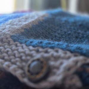 Op de afbeelding: Close-up van een handgebreid item met een knoopsluiting. Het item heeft een patroon van blauwe, grijze en witte strepen. De knoop is donkergrijs met een goudkleurige rand. De gebreide textuur is zichtbaar.