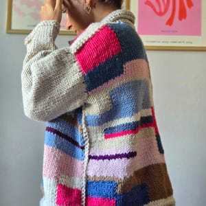 Op de afbeelding: Een handgemaakt vest in patchwork-stijl in verschillende kleuren. De trui heeft grote blokken roze, blauw, bruin en grijs breiwerk. De kraag en manchetten zijn effen grijs.