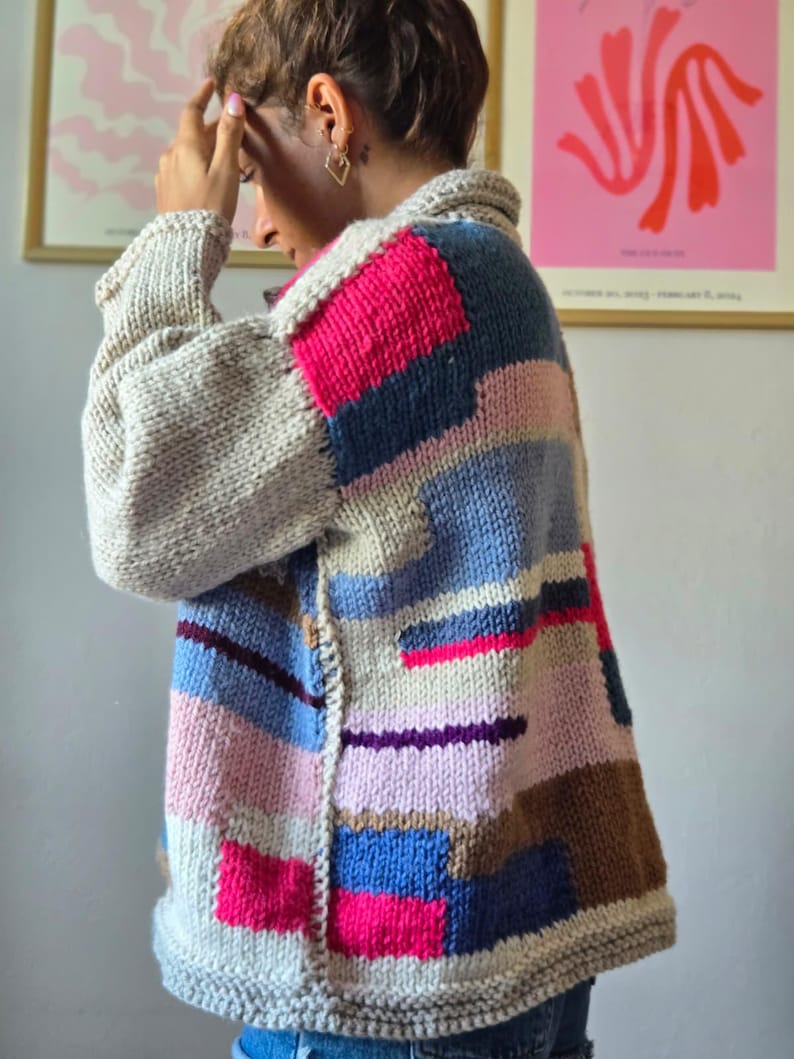 Op de afbeelding: Een handgemaakt vest in patchwork-stijl in verschillende kleuren. De trui heeft grote blokken roze, blauw, bruin en grijs breiwerk. De kraag en manchetten zijn effen grijs.