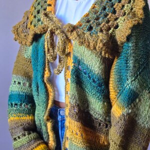 May include: A handmade, open-front cardigan in shades of green, yellow, and blue. The crochet sweater features a unique collar and tie closure. The long sleeves and body have a striped pattern. The garment is made of a thick, textured yarn.