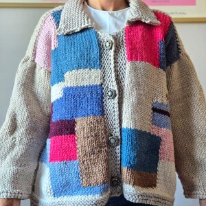 Op de afbeelding: Een grofgebreid vest in een patchwork-ontwerp. De trui heeft een beige basis met kleurrijke vierkanten in roze, blauw, bruin en grijs. Het heeft een kraag, lange mouwen en wordt gesloten met zilveren knopen. Een gezellig, handgemaakt kledingstuk.