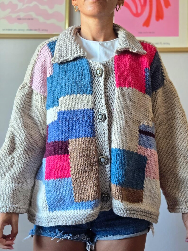 Op de afbeelding: Een grofgebreid vest in een patchwork-ontwerp. De trui heeft een beige basis met kleurrijke vierkanten in roze, blauw, bruin en grijs. Het heeft een kraag, lange mouwen en wordt gesloten met zilveren knopen. Een gezellig, handgemaakt kledingstuk.