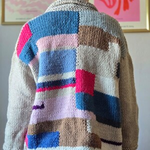 Op de afbeelding: Een dik gebreid vest in een patchwork-ontwerp. De trui heeft een mix van kleuren, waaronder beige, blauw, roze en bruin. De kraag en mouwen zijn effen beige van kleur. De achterkant van de trui is zichtbaar.