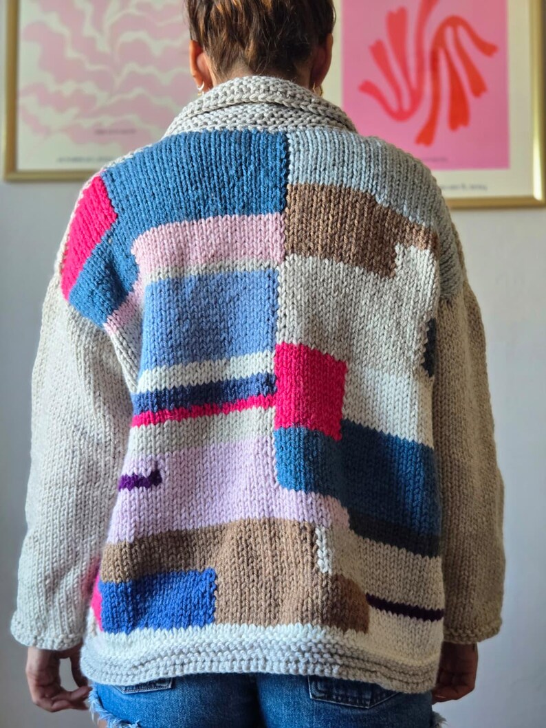 Op de afbeelding: Een dik gebreid vest in een patchwork-ontwerp. De trui heeft een mix van kleuren, waaronder beige, blauw, roze en bruin. De kraag en mouwen zijn effen beige van kleur. De achterkant van de trui is zichtbaar.