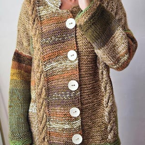 May include: A hand-knitted cardigan in shades of brown, green, and orange. The button-down sweater features a cable knit pattern on the front and sleeves, with large white buttons. The cuffs and collar are trimmed with green yarn.