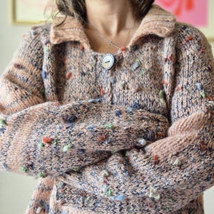 May include: A peach-coloured, button-down cardigan sweater with a collar. The sweater is knitted with a textured pattern and features colourful, raised yarn details. The sleeves are long and the sweater is buttoned up.