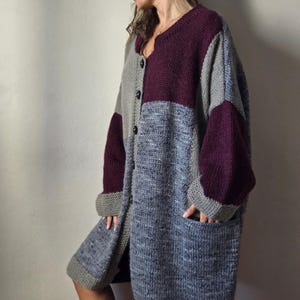 Puede incluir: Un c&aacute;rdigan largo y extragrande con un dise&ntilde;o de patchwork. El su&eacute;ter presenta un patr&oacute;n de bloques de color con secciones grises, azules y burdeos. Tiene cuello en V, mangas largas y dos bolsillos delanteros. El c&aacute;rdigan est&aacute; abotonado y parece estar hecho de un material de punto grueso.