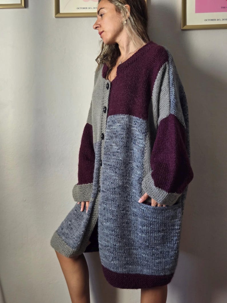 Puede incluir: Un c&aacute;rdigan largo y extragrande con un dise&ntilde;o de patchwork. El su&eacute;ter presenta un patr&oacute;n de bloques de color con secciones grises, azules y burdeos. Tiene cuello en V, mangas largas y dos bolsillos delanteros. El c&aacute;rdigan est&aacute; abotonado y parece estar hecho de un material de punto grueso.