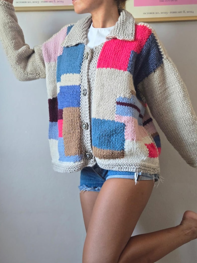 Op de afbeelding: Een kleurrijk patchwork gebreid vest met een kraag en knoopsluiting. De trui heeft een mix van beige, roze, blauwe en bruine vierkanten. De mouwen zijn lang en de stijl is casual en comfortabel.
