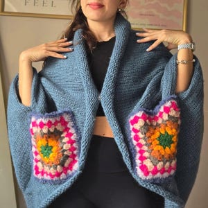 May include: A blue knitted shawl with two square pockets. Each pocket features a colorful, crocheted granny square design with pink, orange, green, and gray accents. The shawl has a wide collar and a cozy, oversized fit.