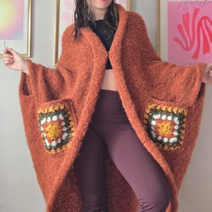 May include: An orange hooded cape with two square pockets featuring a crochet design in yellow, green, and white. The cape is made of a thick, textured material. The model is wearing dark purple pants.