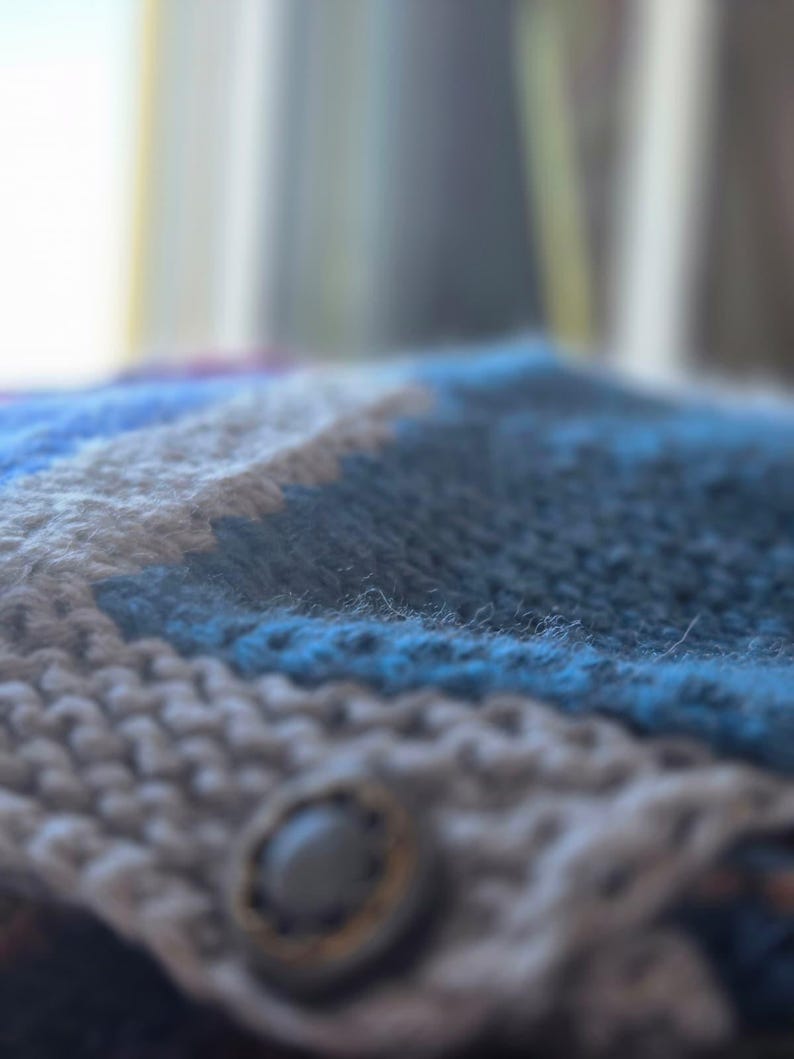 Op de afbeelding: Close-up van een handgebreid item met een knoopsluiting. Het item heeft een patroon van blauwe, grijze en witte strepen. De knoop is donkergrijs met een goudkleurige rand. De gebreide textuur is zichtbaar.