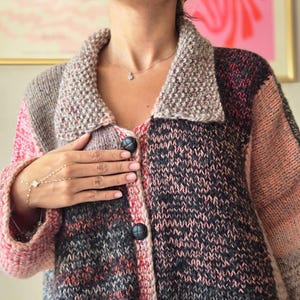 May include: A multi-colored knitted cardigan with a textured collar and large black buttons. The color blocks include shades of pink, gray, and black. The cardigan is a cozy, handmade garment.