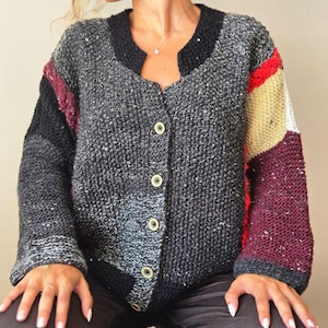 Könnte beinhalten: Knopf-Cardigan in Grau-, Schwarz-, Rot- und Beigetönen. Der Pullover hat ein strukturiertes Strickdesign mit rundem Ausschnitt und langen Ärmeln. Die Ärmel haben farbblockierte Einsätze. Der Cardigan wird mit runden, hellen Knöpfen geschlossen.
