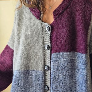 Puede incluir: Un c&aacute;rdigan con botones y bloques de color en tonos grises, morados y azules. El su&eacute;ter de punto presenta un cuello redondo y mangas largas. Los botones son oscuros y el estilo general es acogedor y hecho a mano.