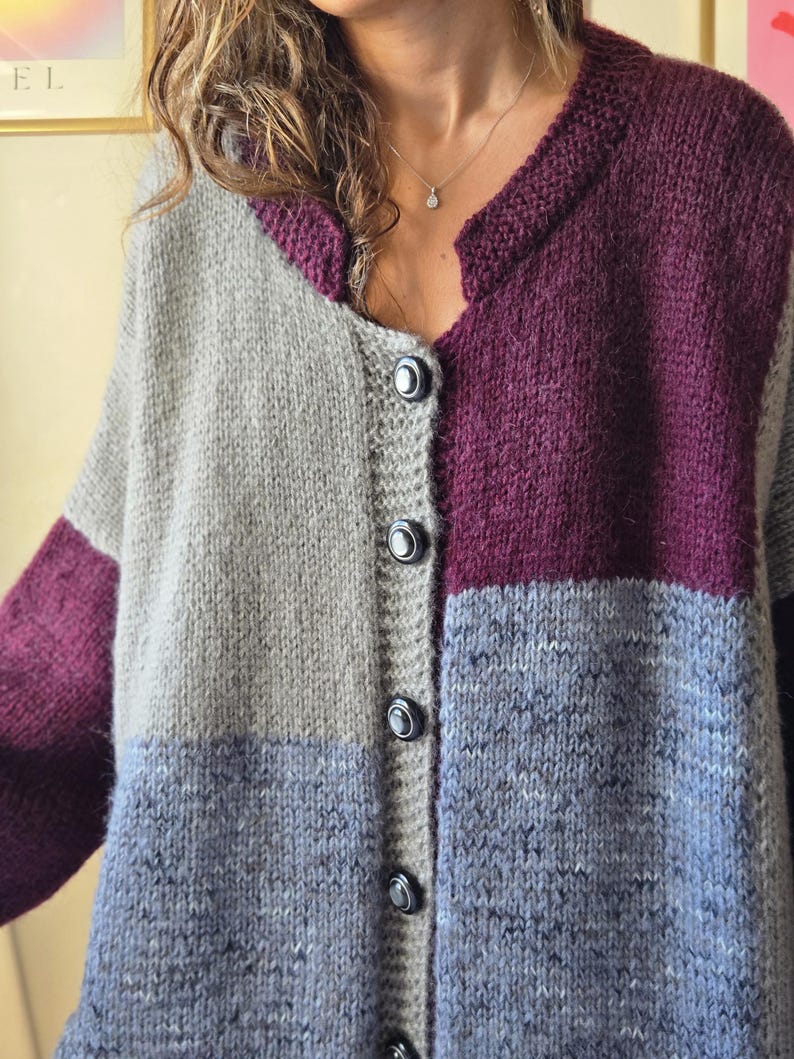 Puede incluir: Un c&aacute;rdigan con botones y bloques de color en tonos grises, morados y azules. El su&eacute;ter de punto presenta un cuello redondo y mangas largas. Los botones son oscuros y el estilo general es acogedor y hecho a mano.