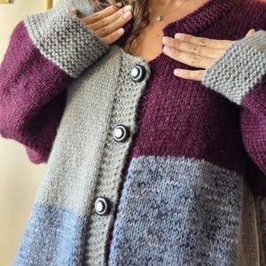 Puede incluir: Un c&aacute;rdigan de punto con bloques de color en tonos grises, morados y azules. El su&eacute;ter tiene cuello en V, mangas largas y grandes botones negros en la parte delantera. Los pu&ntilde;os y la parte superior de las mangas son grises.