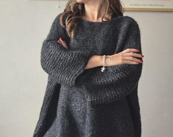 May include: A dark gray, oversized knit sweater with wide sleeves. The sweater has a loose, comfortable fit and a textured knit pattern. The model is wearing black leggings. Two framed art prints are visible in the background.