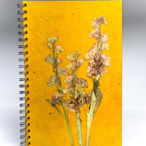 Könnte beinhalten: Ein spiralgebundenes Notizbuch mit einem leuchtend gelben Einband. Der Einband zeigt gepresste, getrocknete Blumen in Rosa- und Weißtönen. Die Spiralbindung des Notizbuchs ist silberfarben.