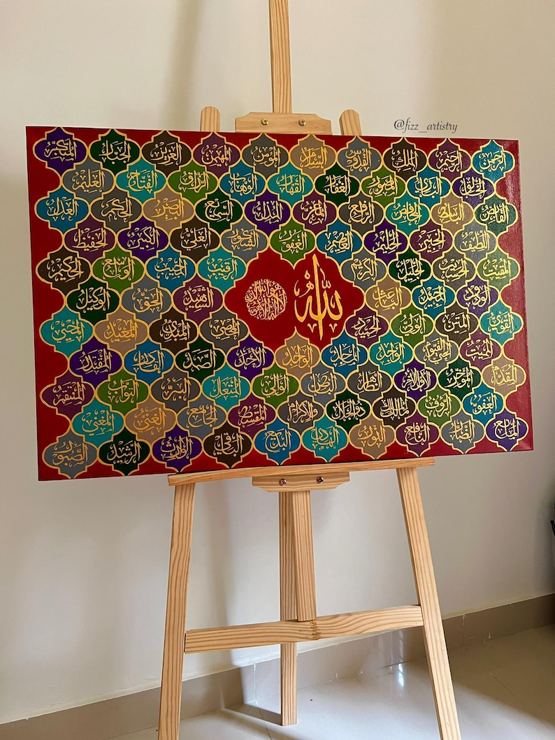 May include: A rectangular canvas art piece featuring Arabic calligraphy in gold lettering on a red background. The artwork is displayed on a wooden easel. The calligraphy is arranged in a grid pattern with various colors.