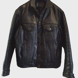 Puede incluir: Chaqueta de cuero negra con cierre de botones, dos bolsillos en el pecho y dos bolsillos laterales. La chaqueta tiene un cuello clásico y mangas largas con puños abotonados. La chaqueta está colgada de una percha negra.