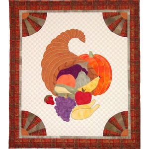 Könnte beinhalten: Ein rechteckiger Quilt mit einem zentralen Füllhorn, das mit einem Kürbis, einem Apfel, Weintrauben und anderen Früchten gefüllt ist. Der Quilt hat einen cremefarbenen Hintergrund mit Rautenmuster und einen rot-braunen Karorand.