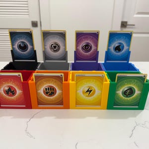 May include: A multi-colored card holder with eight compartments, each holding a trading card. The holder is divided into black, red, orange, yellow, green, blue, purple, and gray sections. The cards feature energy symbols.