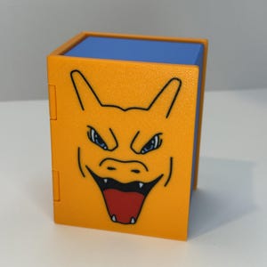 May include: An orange and blue cube-shaped box featuring a cartoon illustration of a dragon's face. The dragon has blue eyes, a red mouth, and sharp teeth. The box is likely a collectible or storage container.