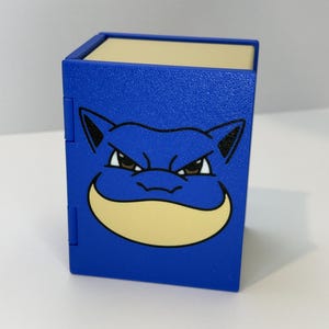 May include: A blue box featuring a cartoon character's face. The character has a light yellow mouth and a stern expression. The box is a solid blue color with black accents.