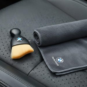 Puede incluir: Un cepillo negro para detallar automóviles con un cabezal de cepillo marrón claro y un mango negro. Una toalla de microfibra gris enrollada y una toalla de microfibra gris doblada con la palabra "Performance" y un logotipo también están en la imagen.