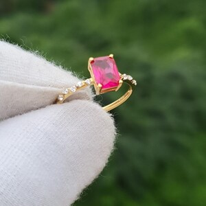 Op de afbeelding: Een gouden ring met een rechthoekige, levendige roze edelsteen. De band van de ring is versierd met kleine, heldere accentstenen. De ring wordt tegen een vage groene achtergrond gehouden, waardoor het ontwerp en de kleur van het sieraad worden benadrukt.