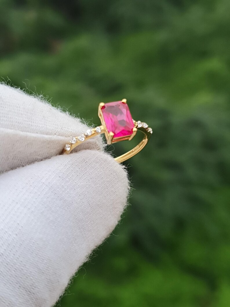Op de afbeelding: Een gouden ring met een rechthoekige, levendige roze edelsteen. De band van de ring is versierd met kleine, heldere accentstenen. De ring wordt tegen een vage groene achtergrond gehouden, waardoor het ontwerp en de kleur van het sieraad worden benadrukt.