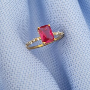 Op de afbeelding: Een gouden ring met een grote, rechthoekige rode edelsteen als middelpunt. De band is ook versierd met kleine, heldere edelstenen. De ring ligt op een lichtblauwe stoffen achtergrond.