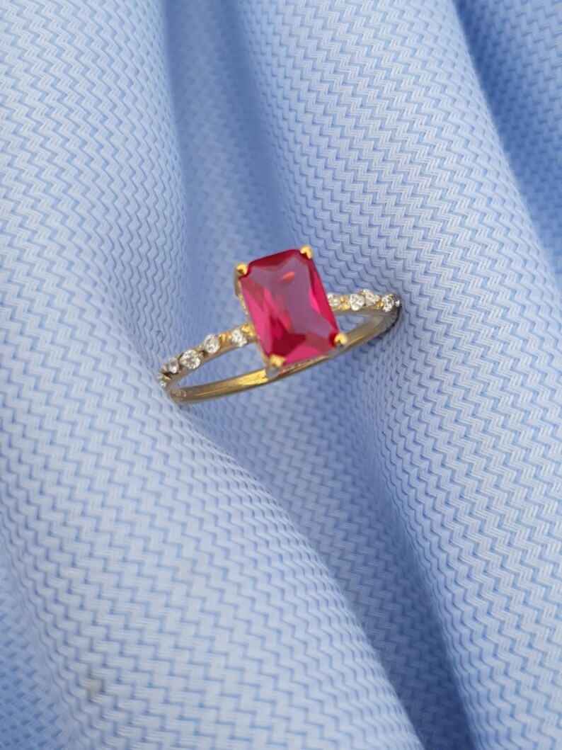 Op de afbeelding: Een gouden ring met een grote, rechthoekige rode edelsteen als middelpunt. De band is ook versierd met kleine, heldere edelstenen. De ring ligt op een lichtblauwe stoffen achtergrond.