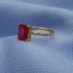 Op de afbeelding: Een gouden ring met een grote, rechthoekige, rode edelsteen als middelpunt. De band is ook goudkleurig, met kleine, heldere edelstenen langs de bovenkant. De ring is geplaatst tegen een lichtblauwe stoffen achtergrond.