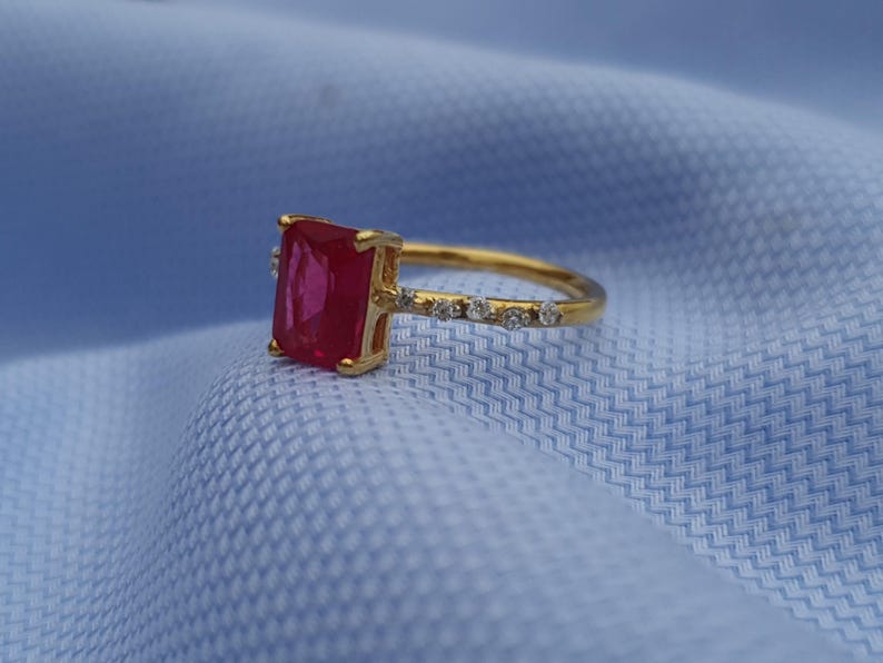 Op de afbeelding: Een gouden ring met een grote, rechthoekige, rode edelsteen als middelpunt. De band is ook goudkleurig, met kleine, heldere edelstenen langs de bovenkant. De ring is geplaatst tegen een lichtblauwe stoffen achtergrond.