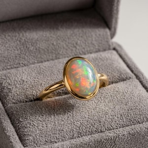 Puede incluir: Un anillo de oro con una piedra preciosa de ópalo cabujón de forma ovalada y lisa. El ópalo muestra un juego de colores con destellos rojos, verdes y naranjas. La piedra está engastada en un bisel de oro pulido sobre una banda fina.